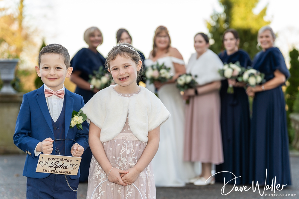 A young boy in a blue suit and a young girl in a pink dress hold a sign saying "don't worry ladies, I'm still single" at the Bagden Hall wedding, with bridesmaids