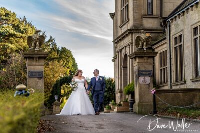 A newlywed couple, Joanne and Brian, walk hand in hand along a path flanked by stone pillars topped with statues, outside a majestic estate, under a clear blue sky.
