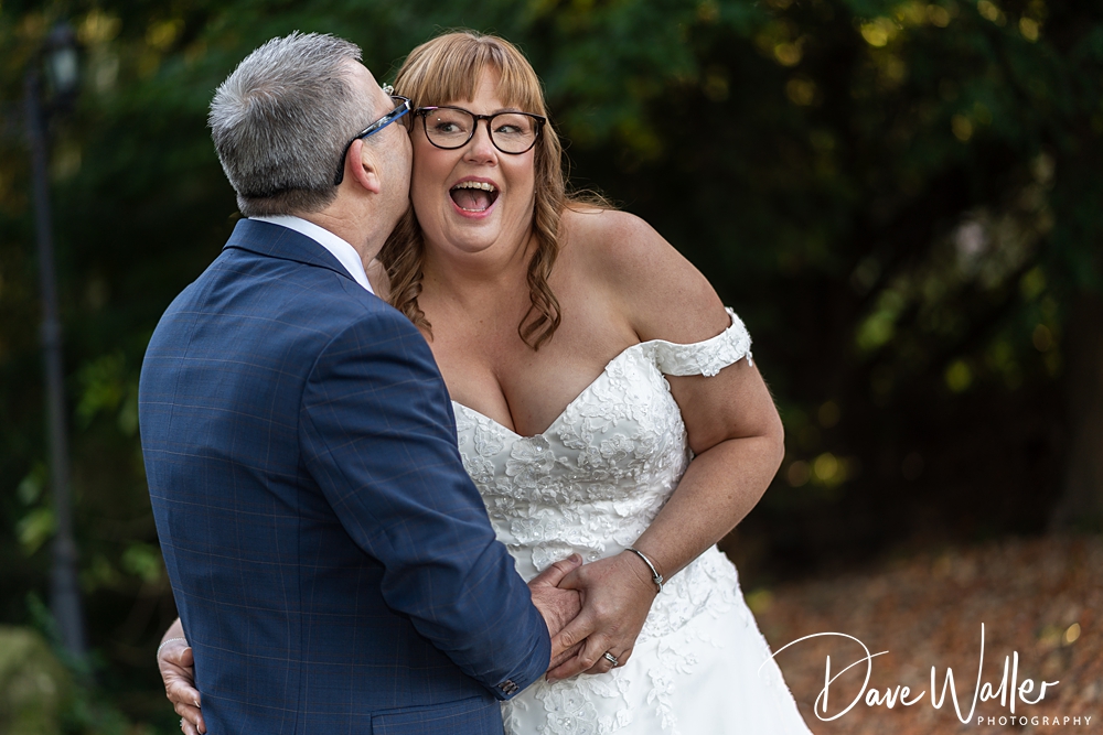 JOANNE and BRIAN, a joyful bride and groom, share a laugh during their wedding photo shoot at Bagden Hall, surrounded by blurred greenery. The bride in a white off-
