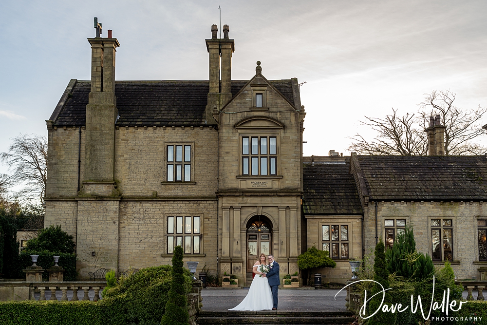 A newlywed couple, JOANNE & BRIAN, embracing in front of an elegant, historic stone building with large windows and chimneys under a clear sky, surrounded by manicured greenery