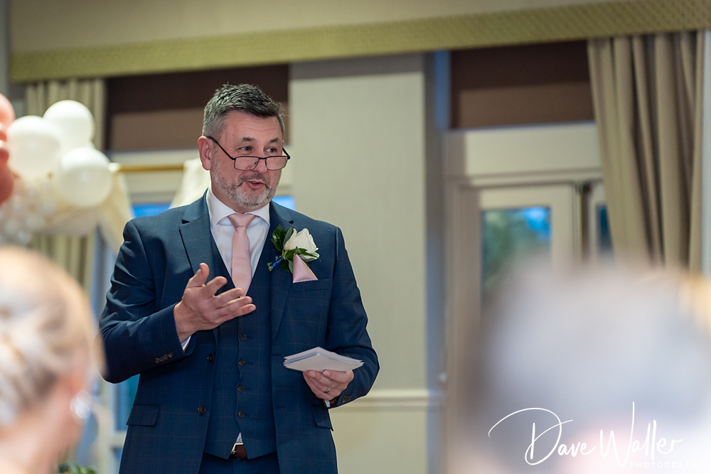 Dave Waller, in a suit with a boutonniere, gives a speech at a wedding, holding a piece of paper. He stands indoors with balloons in the background, addressing an unseen audience