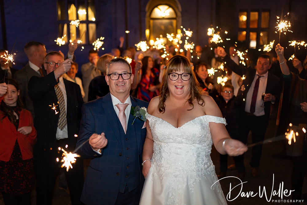 A joyful bride and groom walk through a sparkling tunnel of friends holding lit sparklers at night, expressing happiness and celebration at their Bagden Hall wedding.
