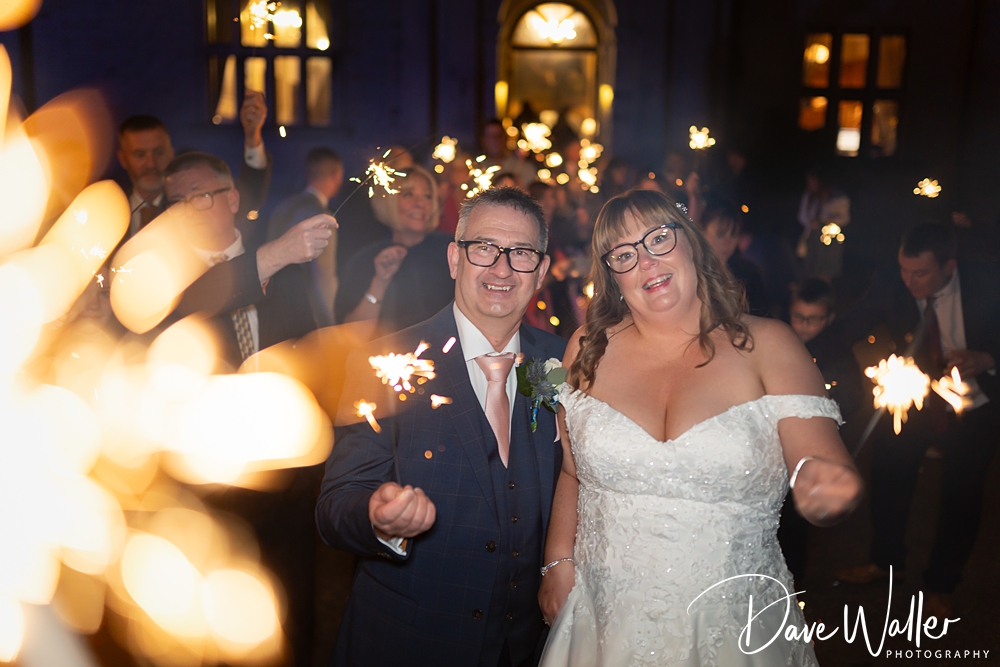 A joyful bride and groom walk hand-in-hand through a sparkler send-off at night, captured by wedding photographer Dave Waller, surrounded by guests, with blurred lights in the foreground adding a magical feel