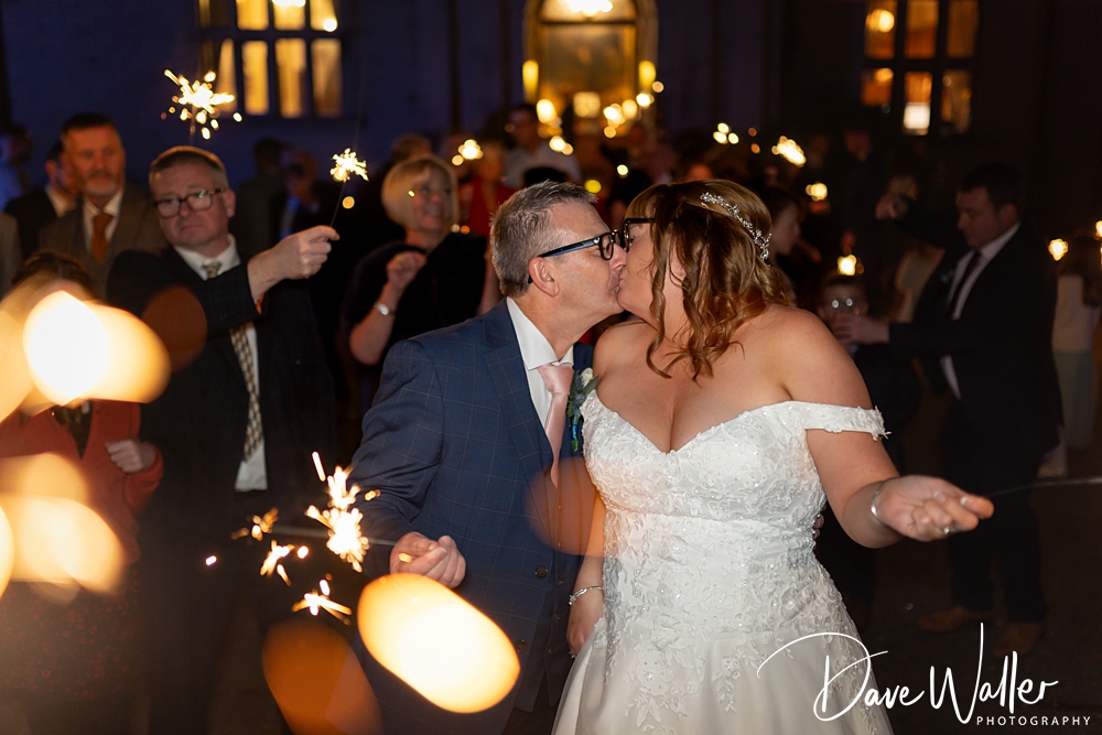 A newlywed couple, JOANNE & BRIAN, kisses amidst a sparkling crowd holding sparklers at their Bagden Hall evening wedding reception, warmly illuminated by twinkling lights.