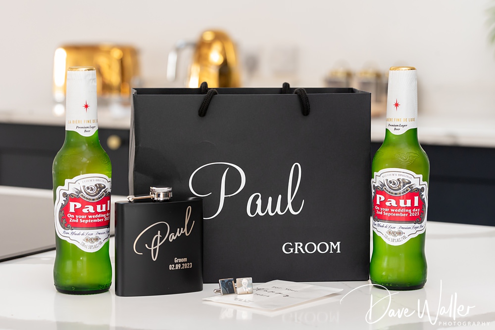 A black gift bag labeled "Paul Groom" is flanked by two bottles of beer. In front of the bag are a black flask, engraved with "Paul" and a date, a pair of cufflinks, and a card. The image, capturing the elegant setting at Woodlands Hotel Leeds, is signed "Dave Walker Photography".