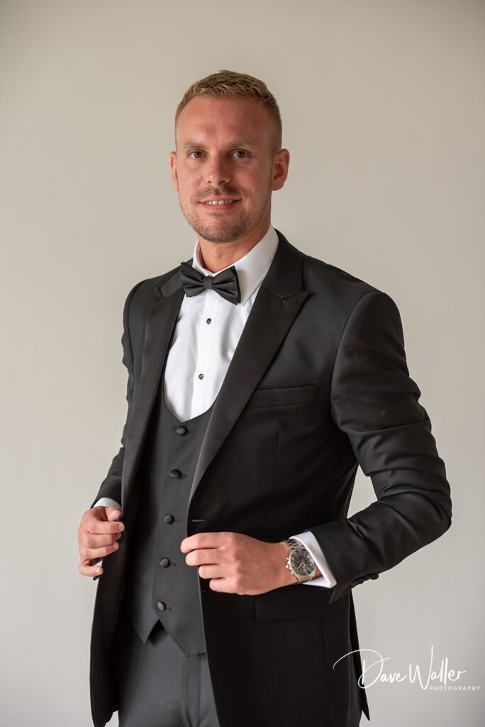 A person dressed in a black tuxedo with a bow tie stands against a plain background, holding the lapels of their jacket. A logo in the bottom right corner reads "Dave Waller Photography." This image exudes sophistication, reminiscent of an elegant evening at the Woodlands Hotel Leeds.