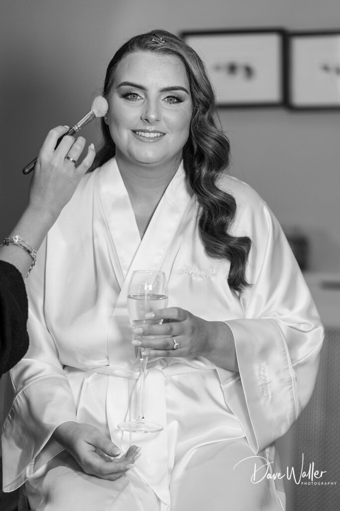 A woman with long, wavy hair sits in a white satin robe at Woodlands Hotel Leeds, holding a glass of champagne. She's smiling while a hand applies makeup to her face with a brush. The image has a watermark on the bottom right corner that reads "Dave Waller Photography.