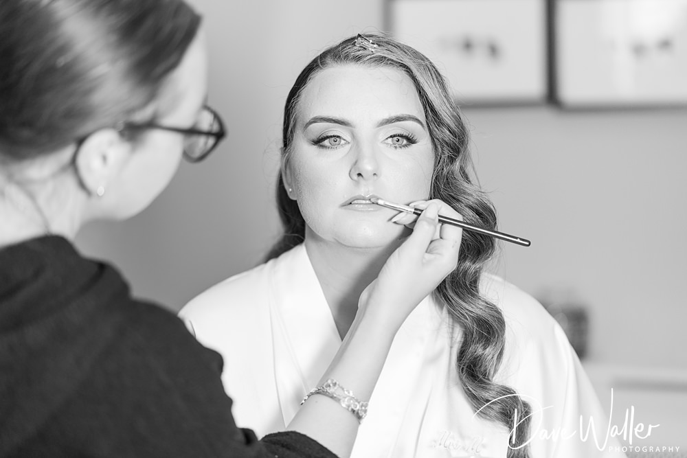 A makeup artist applies lipstick to a woman with wavy hair and a white robe at the Woodlands Hotel Leeds. The woman has a serene expression, and her eyes are focused. The background is softly blurred, with indistinct details. Black and white photo by Dani Waller Photography for Hannah & Paul's wedding.