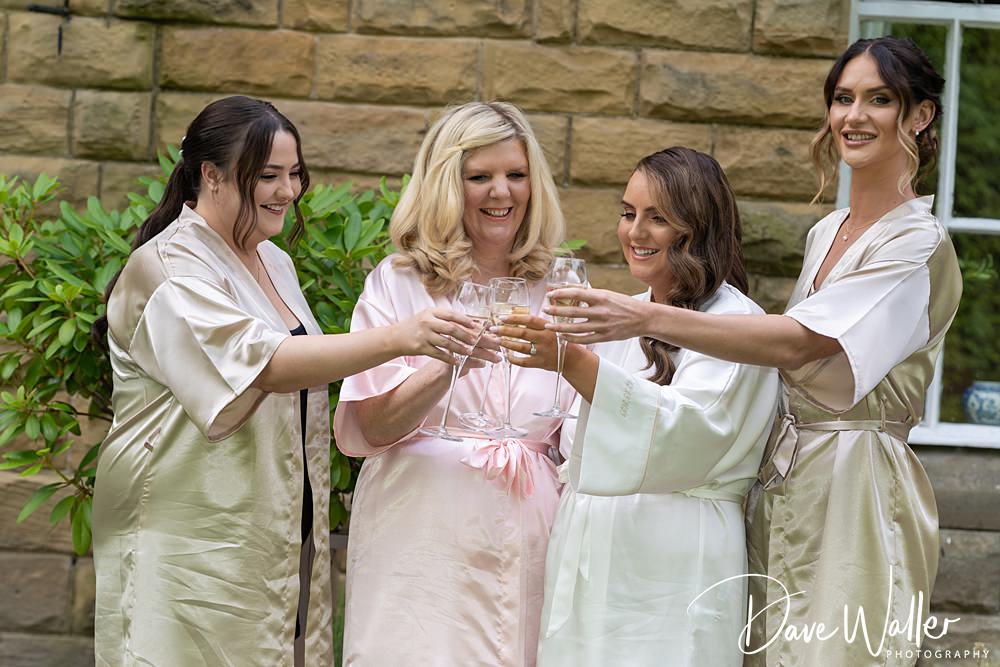 Four women in satin robes toast with champagne flutes outside, smiling and enjoying the moment. The stone wall and greenery at Woodlands Hotel Leeds add to the festive atmosphere, making it a memorable celebration for Hannah & Paul.