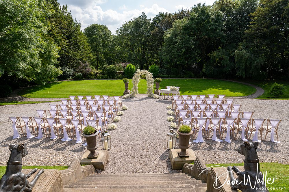 An outdoor wedding ceremony setup at the Woodlands Hotel Leeds features rows of white chairs arranged on a gravel path leading to a floral arch. The scene is surrounded by lush greenery and trees. The sky is partly cloudy, and the setting is at the bottom of a grand staircase with stone balustrades.