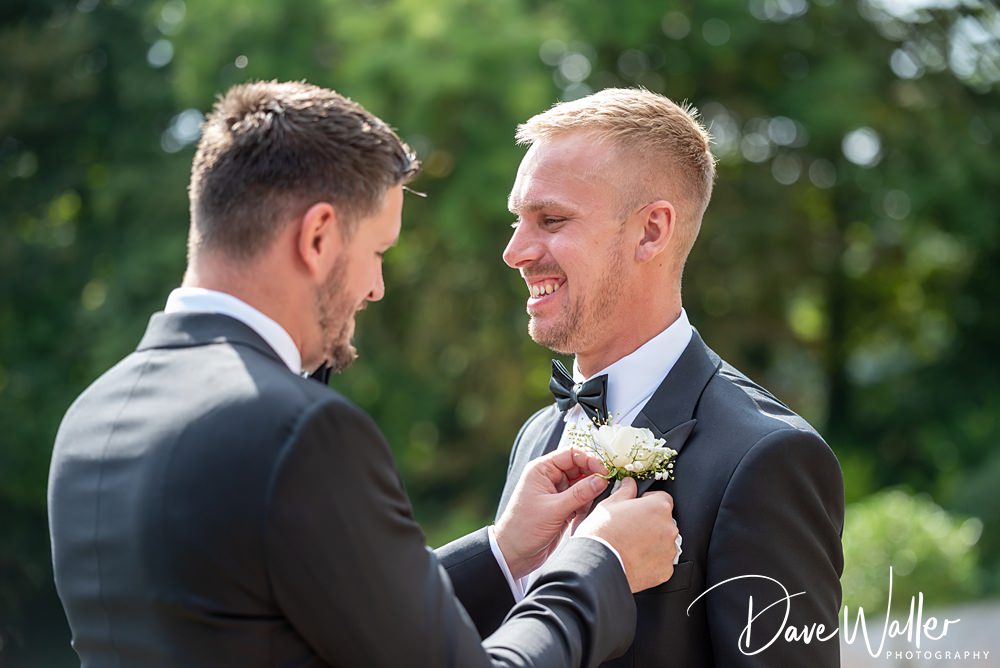 Two men in formal black suits are outside the Woodlands Hotel Leeds on a sunny day. One man is pinning a white boutonniere onto the other's jacket. They are both smiling, with lush greenery blurred in the background. The photo is watermarked with "Dave Waller Photography.