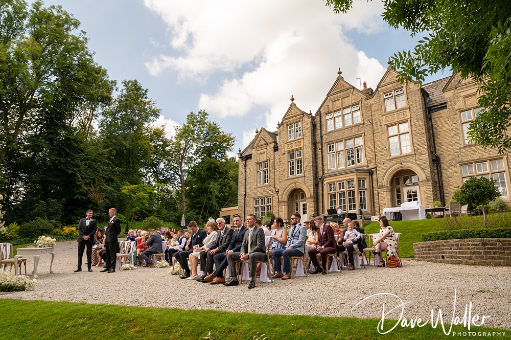 Outdoor wedding ceremony at the Woodlands Hotel Leeds, with guests seated on wooden benches in front of a grand historic building surrounded by trees. Hannah & Paul are standing with the officiant on a stone path, and a few fluffy clouds are visible in the sky.