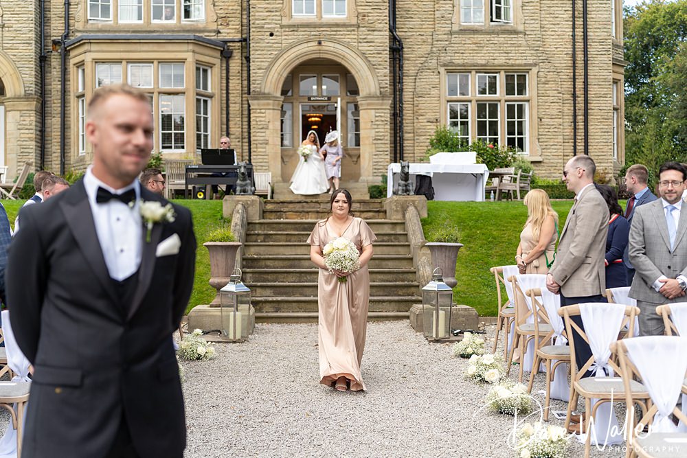 A groom waits at the end of an outdoor aisle at the Woodlands Hotel Leeds as a bridesmaid in a satin dress walks toward him holding a bouquet. Guests are seated on either side of the aisle. In the background, Hannah & Paul descend the steps of this historic Leeds hotel, ready to exchange vows.