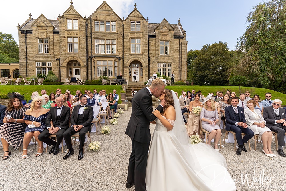 At Woodlands Hotel Leeds, Hannah and Paul share a kiss at their outdoor wedding ceremony in front of a grand, historic building. Guests are seated in rows, smiling and watching the couple. The day appears sunny, with clear skies and a well-maintained lawn in the background.