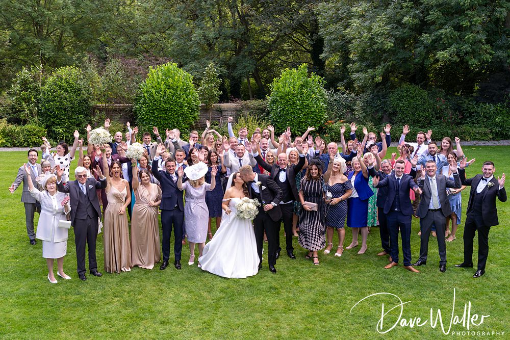 A large group of well-dressed people, including Hannah & Paul in the foreground, pose outdoors on a lush green lawn at Woodlands Hotel Leeds. Many guests are smiling, waving, and raising their hands in celebration. Trees and bushes form a backdrop.