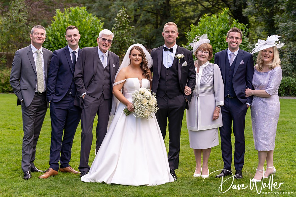 A wedding party of seven stands outdoors on the lush lawn of Woodlands Hotel Leeds. The bride, in a white gown holding a bouquet, stands beside the groom in a black tuxedo. The others are dressed in formal attire, including suits and dresses. Trees and greenery provide a picturesque backdrop.