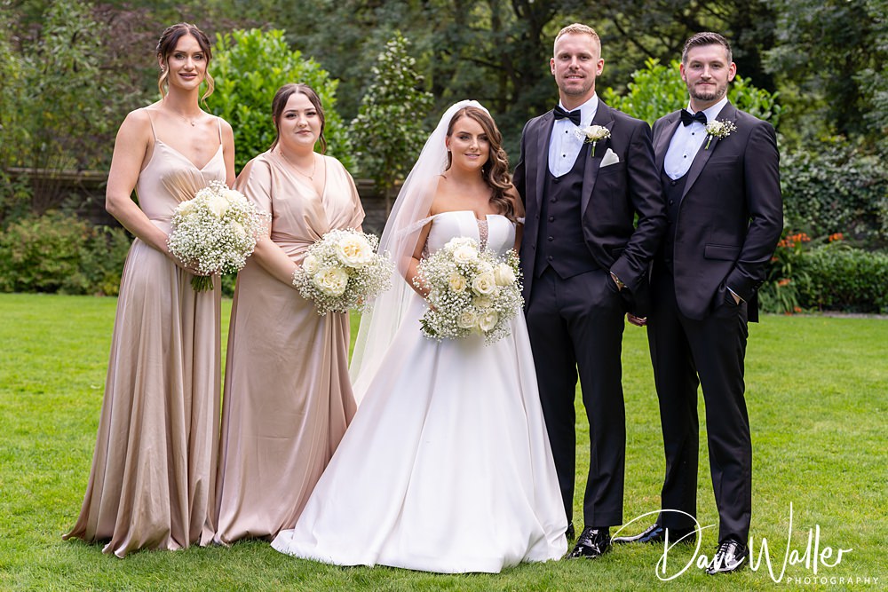 A wedding party stands on the grass with trees in the background at Woodlands Hotel Leeds. The bride, Hannah, is in a white gown and veil, holding a bouquet, with two bridesmaids in beige dresses to her left. Two groomsmen in black tuxedos stand to her right. Everyone is looking at the camera.