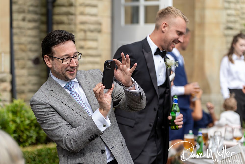 A man in a grey checkered suit and glasses is taking a photo with his phone. Behind him, another man in a black tuxedo with a boutonnière is holding a beer bottle and smiling. They are outdoors near the Woodlands Hotel Leeds, by a stone building, with people mingling in the background.