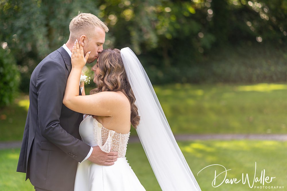 A couple shares a tender kiss on their wedding day at Woodlands Hotel Leeds. The groom, in a black suit, gently holds the bride's face, while the bride, in a white wedding dress and veil, tenderly touches his cheek. They stand on a grassy area with lush greenery in the background.