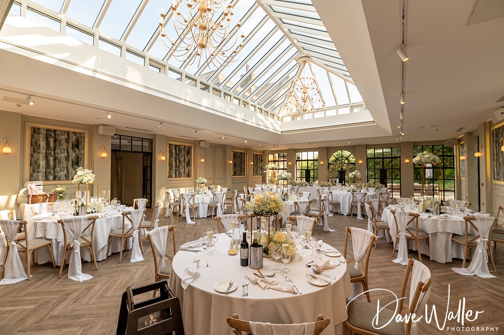 A beautifully decorated wedding reception venue at Woodlands Hotel Leeds with round tables covered in white linens and elegant centerpieces under a glass ceiling. Chandeliers hang from the ceiling, creating a warm ambiance. The room is bright with natural light. Hannah & Paul's photographer's watermark is present.