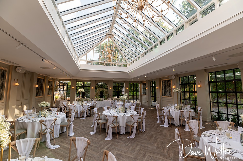 A well-lit, elegant wedding reception hall at the Woodlands Hotel with a glass ceiling and chandeliers. Round tables are set with white tablecloths, floral centerpieces, and wooden chairs with white covers. Large windows offer a view of greenery outside. Photography credit is visible.