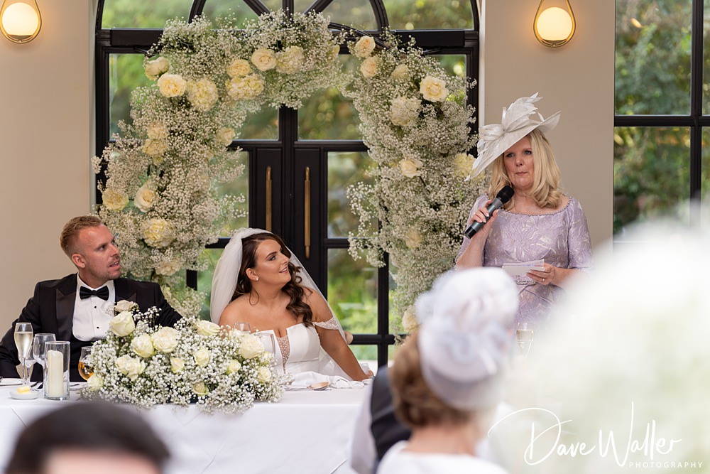 A woman in a lavender dress and hat is speaking into a microphone at the Woodlands Hotel Leeds wedding reception. The bride and groom, seated at a table with a floral arrangement, look on. The table is decorated with white flowers and glasses, with a large floral arch in the background.