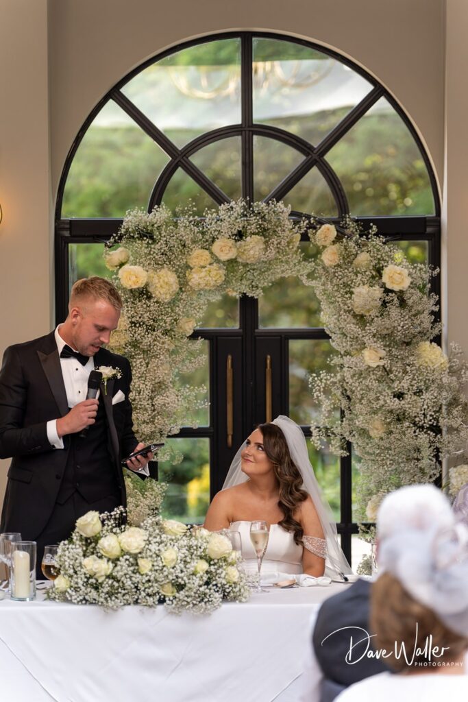 A groom wearing a black tuxedo is giving a speech using a microphone while standing beside a seated bride in a white wedding dress. They are under a floral arch decorated with white flowers and baby's breath at the Woodlands Hotel Leeds. The backdrop is a large arched window, perfect for Hannah & Paul's special day.