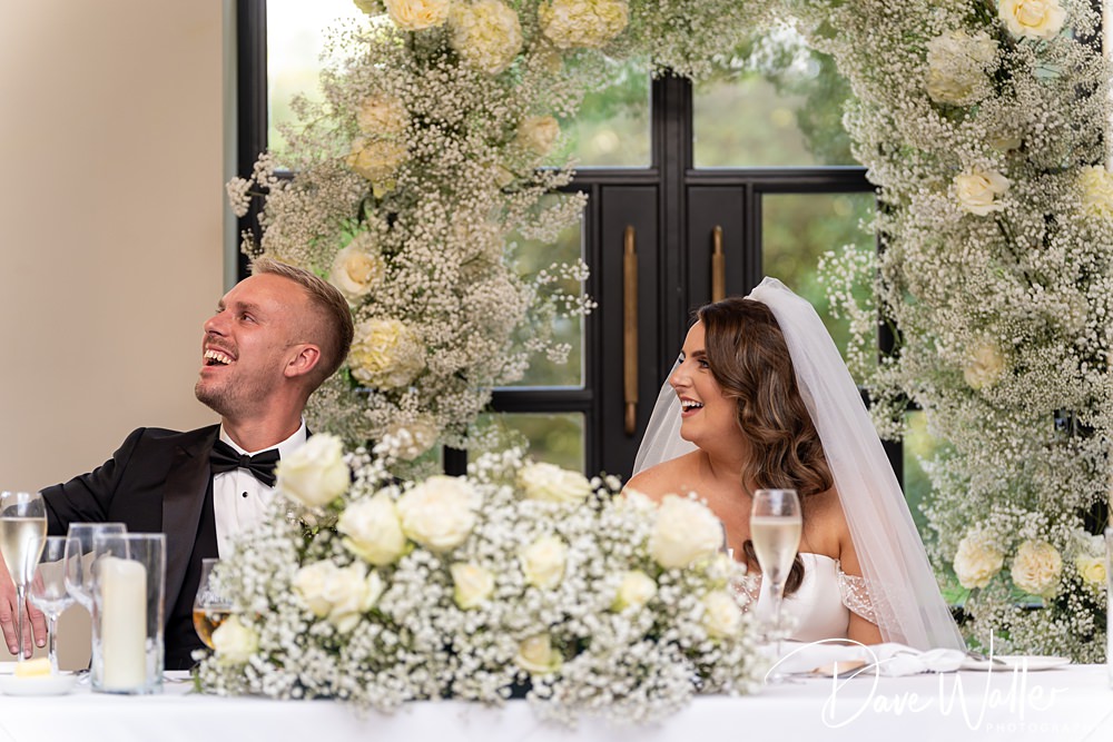 A bride and groom, Hannah and Paul, sit at a table adorned with white roses and baby's breath. Behind them is a large floral arch made of white blooms and greenery at the Woodlands Hotel Leeds. Both are smiling and looking to the side, dressed elegantly for their wedding day.