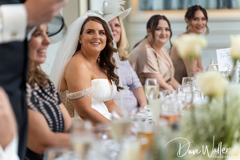 A smiling bride in a white dress and veil sits at a wedding reception table with several guests at the Woodlands Hotel Leeds. She looks towards someone giving a speech. The table is adorned with glasses and flowers. Guests in various attire are seated beside her. Photo credit: "Dave Waller Photography".