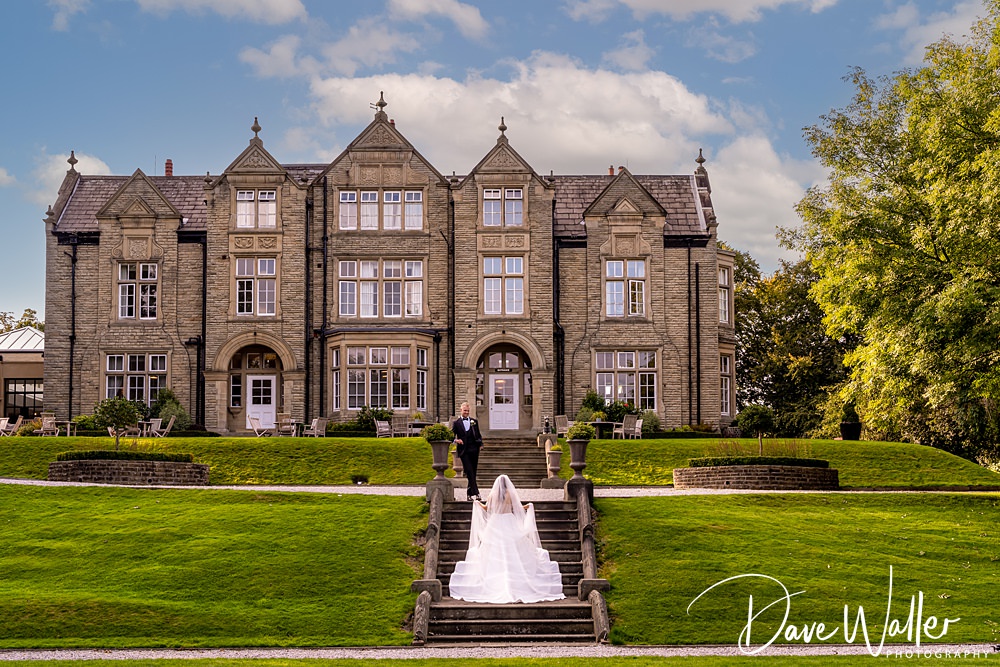 A bride in a white wedding dress walks up stone steps toward a groom on a green lawn, leading to the elegant Woodlands Hotel Leeds with tall, narrow windows and intricate details. The sky is partly cloudy. "Hannah & Paul - Dave Waller Photography" is written in the corner.