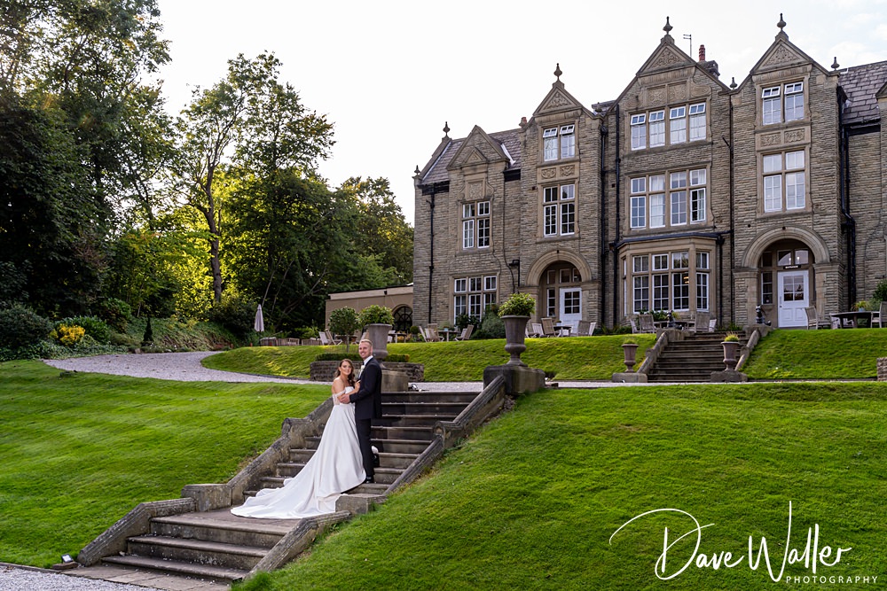 A bride and groom, Hannah & Paul, stand on outdoor steps leading up to the grand Woodlands Hotel Leeds with large windows and manicured lawns. The bride is wearing a long white dress, and the groom is in a dark suit. Lush greenery surrounds the elegant structure. Photographer credit: Dave Waller.