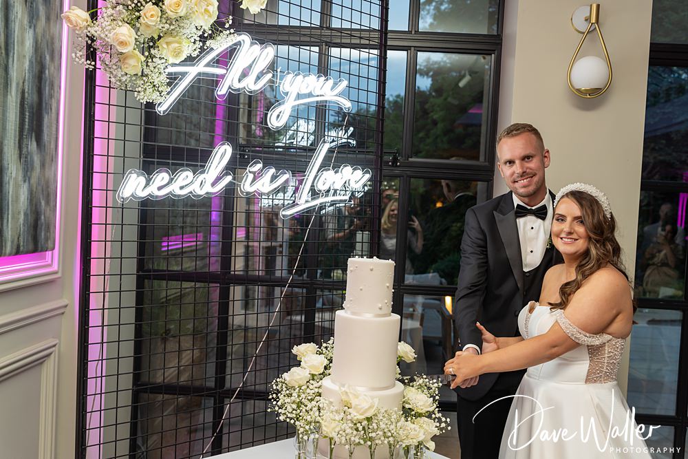 A bride and groom, Hannah & Paul, smile as they cut a white, three-tiered wedding cake adorned with flowers. The backdrop at the Woodlands Hotel features a neon sign that reads "All you need is love." The groom is in a black tuxedo, and the bride wears a white dress and headpiece.