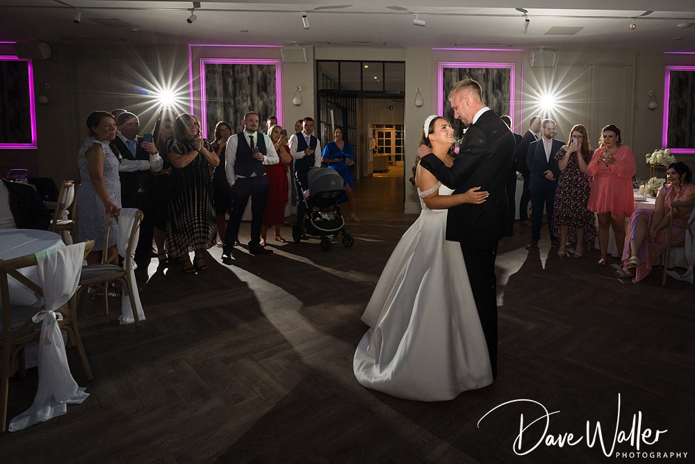 A bride and groom share their first dance in the center of a dimly lit room, surrounded by guests who are watching and smiling. The bride wears a white gown and the groom is in a black suit. The setting at Woodlands Hotel has bright lights in the background with purple accents, perfect for any wedding photographer to capture.