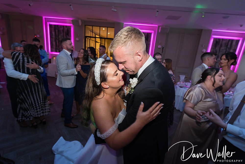 A bride and groom embrace and smile at each other while dancing in a brightly lit reception hall at Woodlands Hotel Leeds. Guests in formal attire enjoy themselves in the background. Neon lights in pink add to the celebratory atmosphere. The image is captured by Dave Waller Photography.