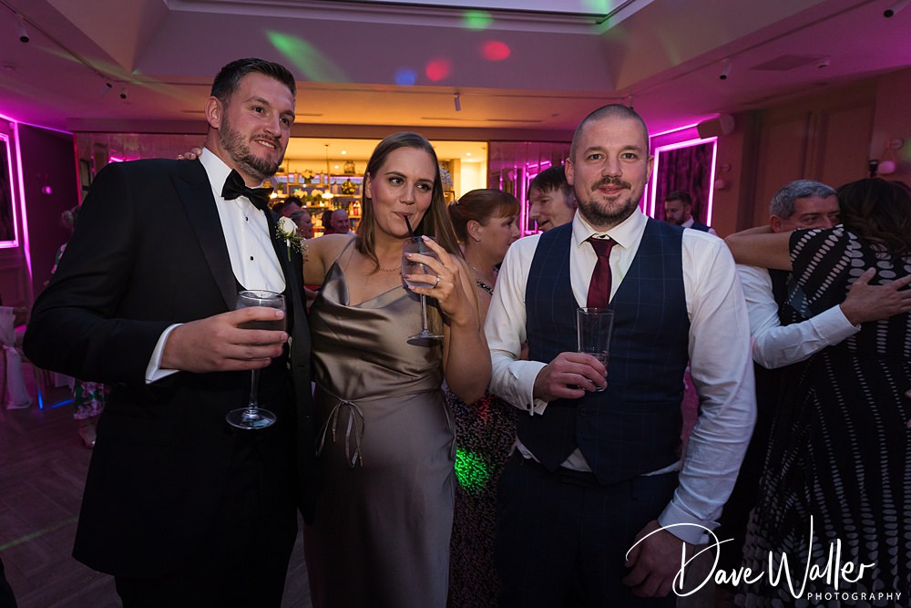 Three people dressed formally are enjoying a party at the Woodlands Hotel Leeds. A man in a tuxedo, a woman in a satin dress, and another man in a suit pose with drinks in hand. Colorful party lights and a crowd in the background can be seen. The image is credited to Dave Waller Photography.