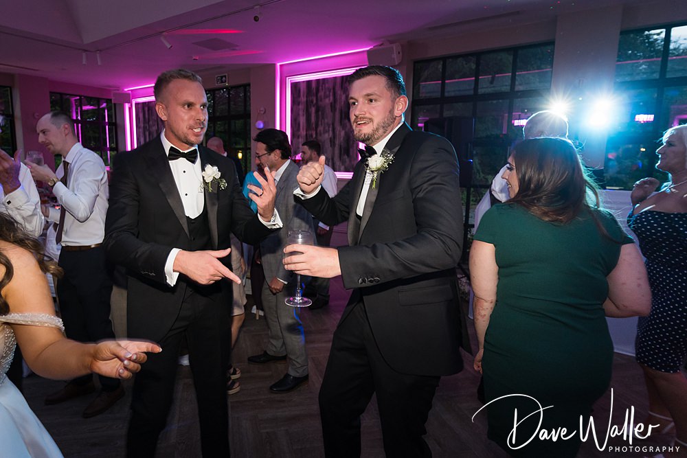 A lively wedding reception at Woodlands Hotel Leeds with two men in tuxedos dancing and pointing fingers at each other. Around them, guests are enjoying Hannah & Paul's celebration. The room is lit with a vibrant pink hue. The photo is by Dave Walker Photography, as indicated by the watermark.