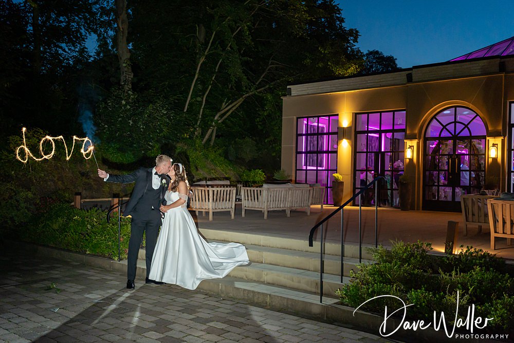 A newlywed couple, Hannah & Paul, wearing formal attire, stands outside the Woodlands Hotel Leeds at night. The groom holds a sparkler, creating the word "love" in the air. The bride, in a white gown, embraces him. The background features trees and a purple-lit Leeds hotel.