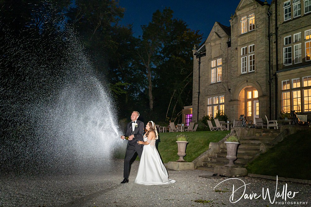Hannah & Paul stand outside the grand Woodlands Hotel at night. The groom, dressed in a tuxedo, sprays what appears to be champagne from a bottle, while the bride, in her wedding gown, watches and smiles. The illuminated building behind them creates a festive scene.