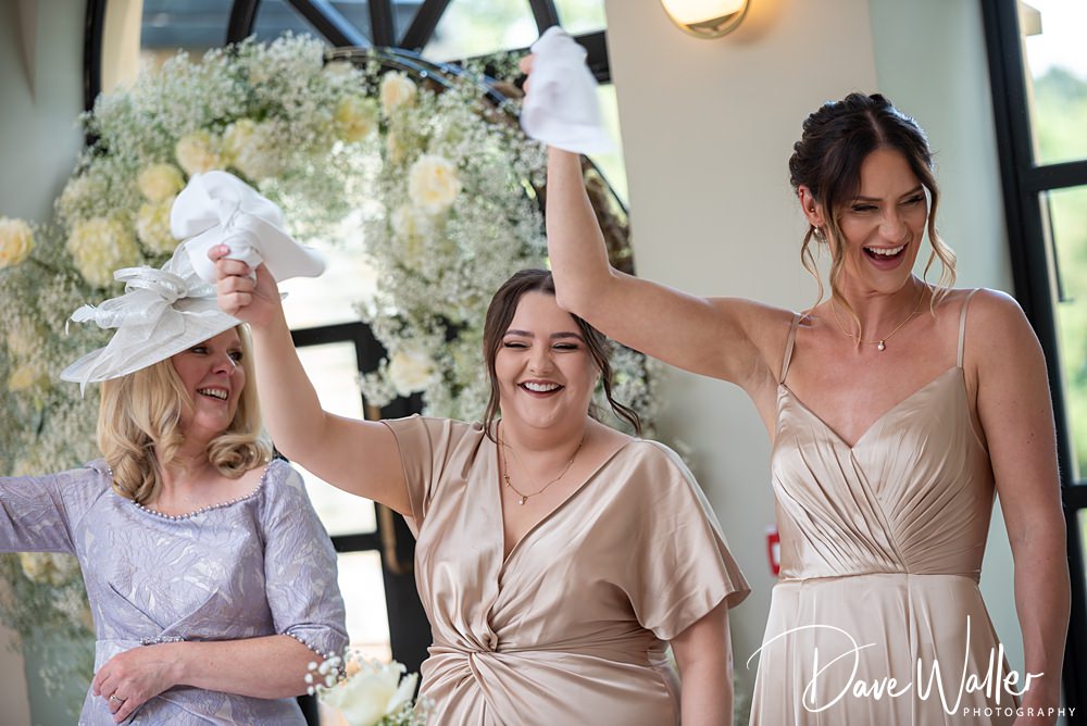 Three women in elegant dresses smile and wave white napkins in the air at Hannah and Paul’s wedding at the Woodlands Hotel Leeds. The left woman wears a light purple dress with a matching hat, the center woman dons a mauve gown, and the right woman is in a champagne-colored dress.