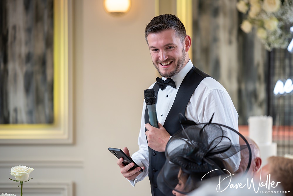 A man wearing a black vest and bow tie is holding a microphone and speaking, while looking at his phone. He appears to be giving a speech at an indoor event at the Woodlands Hotel in Leeds. A woman in a fancy hat, possibly Hannah or Paul’s guest, is seated in front of him, listening attentively.