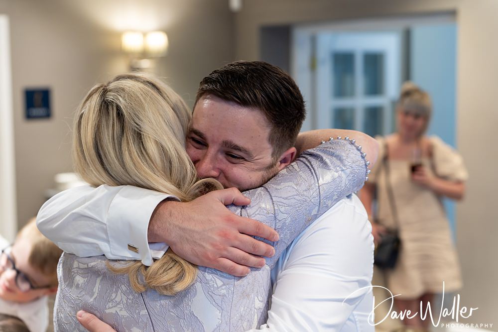 A person with short dark hair and a white shirt warmly embraces another person with long blonde hair and a patterned light blue top. In the softly lit indoor space of Woodlands Hotel, Leeds, another individual in focus holds a phone, capturing the heartfelt moment.