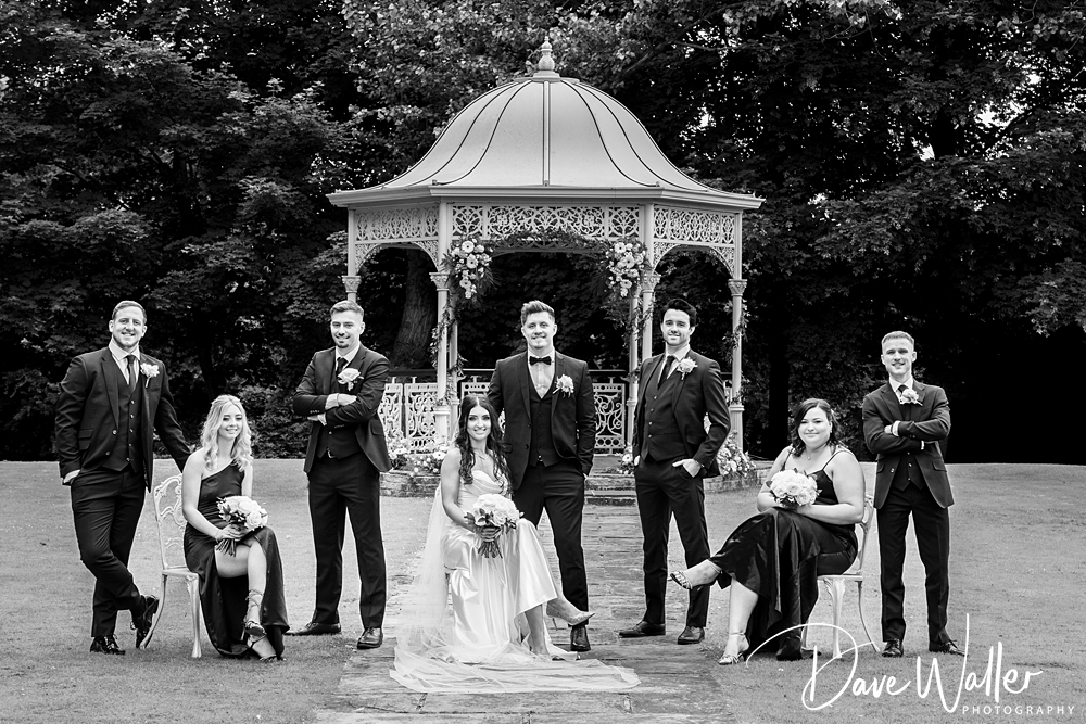 A black-and-white photo shows a wedding party. The bride and groom sit in front of an ornate gazebo at Bagden Hall, surrounded by four groomsmen and two bridesmaids. Everyone is dressed formally and holding bouquets. The lush garden in the background completes the scenic setting, captured perfectly by the wedding photographer.