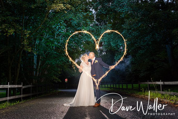 A bride and groom stand on a dimly lit path surrounded by trees at Bagden Hall. They hold sparklers that form a glowing heart shape above their heads. The bride is in a white gown and the groom in a suit. The photographer's signature is in the corner: "Dave Waller Photography.