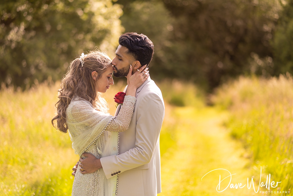 A couple stands in a sunlit, grassy pathway embracing. The man, dressed in a light-colored suit, tenderly kisses the forehead of the woman, who is dressed in an elegant white outfit and holding a red rose. Captured by Bagden Hall Wedding Photographer, they are surrounded by nature, with tall grass and trees in the background.