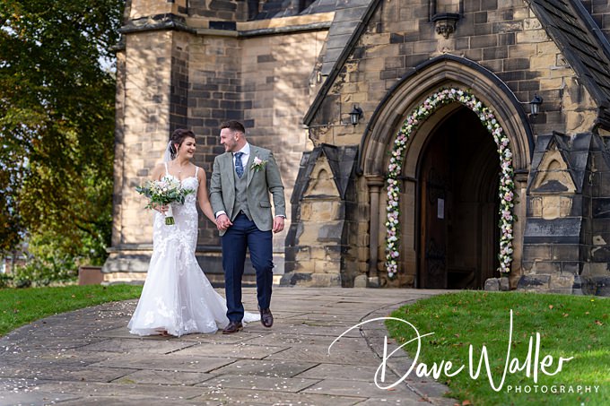 A bride in a white dress and a groom in a gray suit walk hand in hand, smiling at each other, in front of a stone church entrance adorned with flowers. The setting is peaceful with greenery and the path leading to the church. Captured beautifully by Bagden Hall Wedding Photographer, Dave Waller.