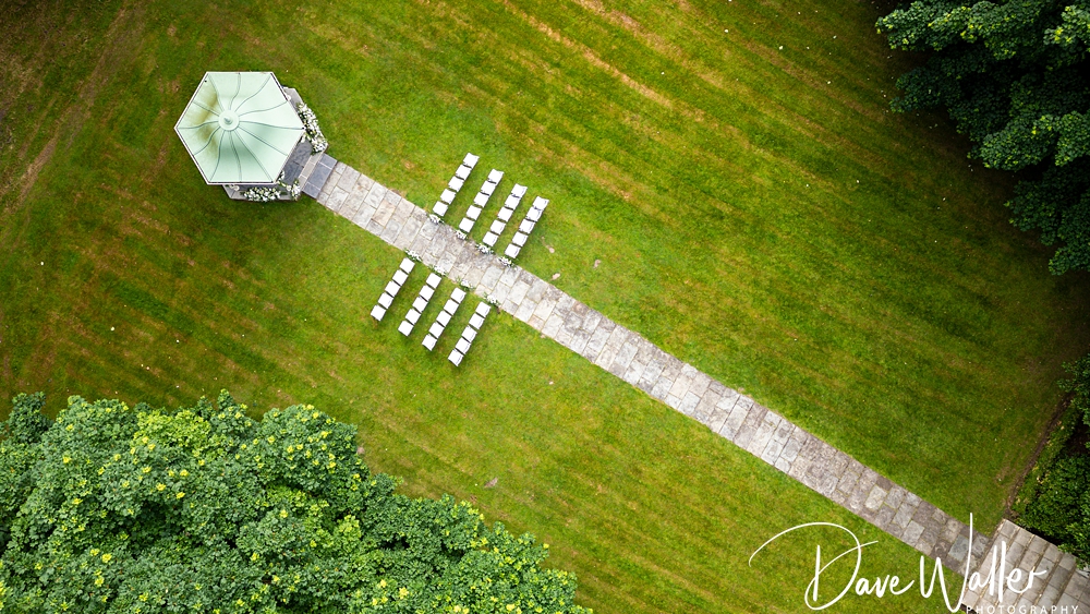 Aerial view of an outdoor setup for a wedding ceremony in a park. Rows of white chairs are arranged along a stone pathway leading to a gazebo, surrounded by green grass and trees. "Bagden Hall Wedding Photographer - Dave Walker Photography" is written in the bottom right corner.