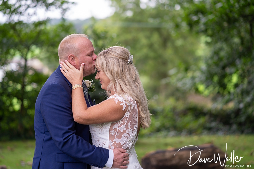 A groom in a navy suit kisses the forehead of a bride in a lace wedding dress as they embrace outdoors, framed by lush green trees and blurred nature. The couple appears to be in a moment of tender affection. Text on the image reads "315 Bar and Restaurant Wedding Photography by Dave Waller.