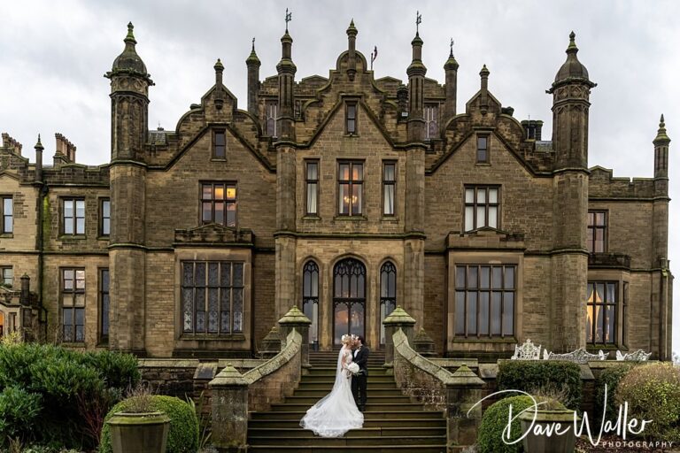 Laura and Jake, in their elegant wedding attire, embrace on the steps of Allerton Castle. The grand historic stone mansion, adorned with many windows and ornate architectural details, stands majestically against a cloudy sky.