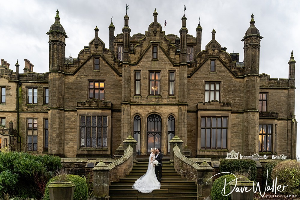 Laura and Jake, in their elegant wedding attire, embrace on the steps of Allerton Castle. The grand historic stone mansion, adorned with many windows and ornate architectural details, stands majestically against a cloudy sky.
