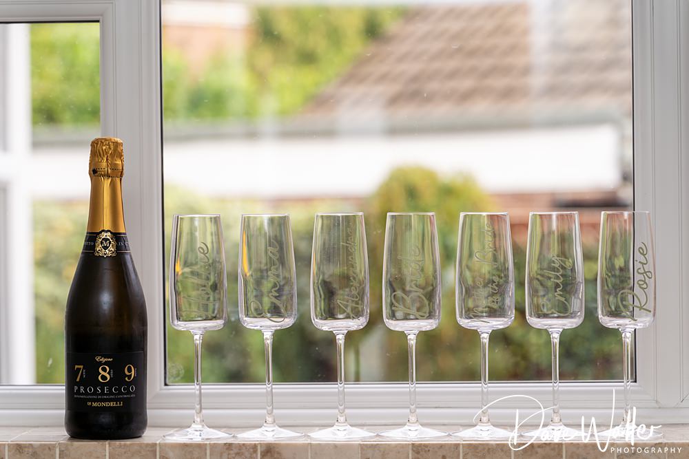 A bottle of 789 Prosecco stands on a windowsill at Wentbridge House beside six empty champagne flutes, each etched with the word "Prosecco." In the background, a blurred view of a garden hints at the elegance of Georgia & Luke's unforgettable wedding.
